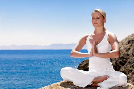 Eine Frau in weißer Kleidung sitzt in Meditationshaltung am Meer, die Hände vor dem Brustkorb gefaltet, was eine aufrechte Haltung und innere Ruhe ausstrahlt.